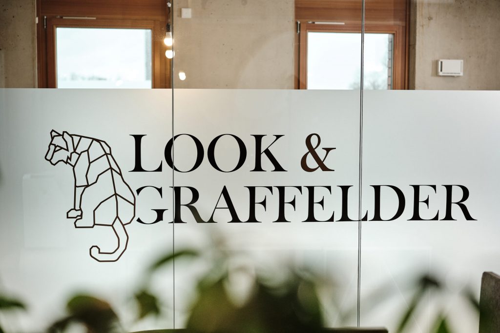 Eine Glaswand zeigt das Logo „Look & Graffelder“ mit einem geometrischen Tiger, im Hintergrund ein modernes Büro mit Pflanzen.
