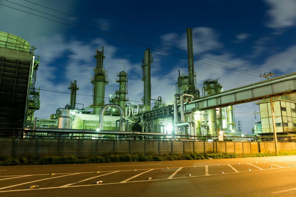 Großindustrielle Anlage mit Rohrleitungen und Schornsteinen bei Nacht. Weitläufiges Gelände, künstliche Beleuchtung, Fokus auf Industriearchitektur.